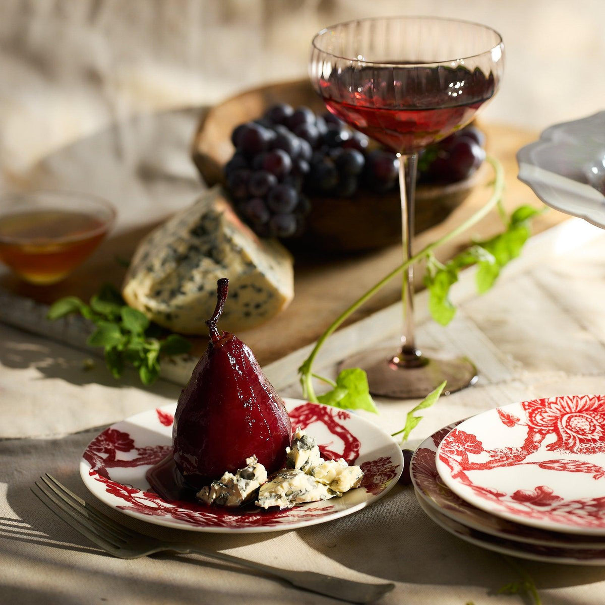 A poached pear elegantly graces the Arcadia Crimson Small Plates from Caskata Artisanal Home, complemented by blue cheese and paired with a glass of red wine. Grapes and an extra piece of cheese complete this captivating tablescape.