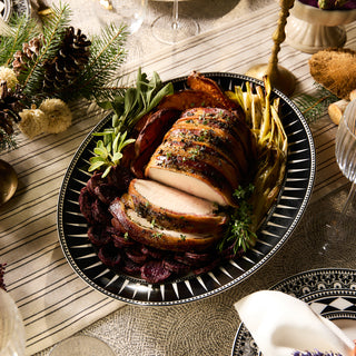 The Marrakech Oval Rimmed Platter by Caskata Artisanal Home features sliced roasted meat garnished with herbs, surrounded by vegetables, elegantly displayed on a patterned tablecloth adorned with candles and pinecones.