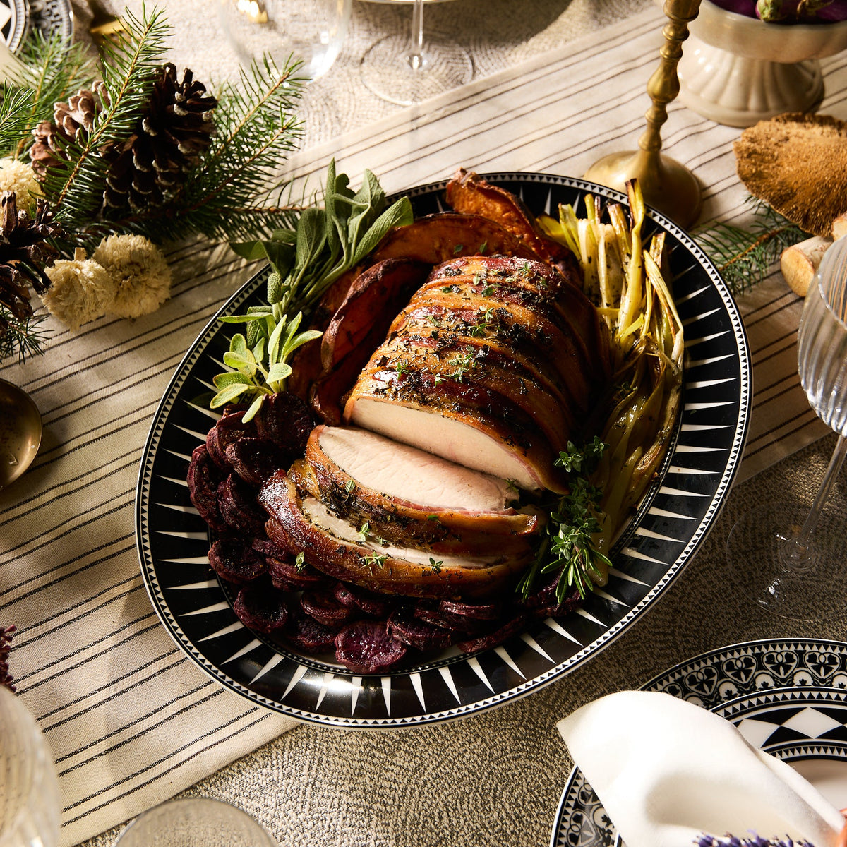 The Marrakech Oval Rimmed Platter by Caskata Artisanal Home features sliced roasted meat garnished with herbs, surrounded by vegetables, elegantly displayed on a patterned tablecloth adorned with candles and pinecones.