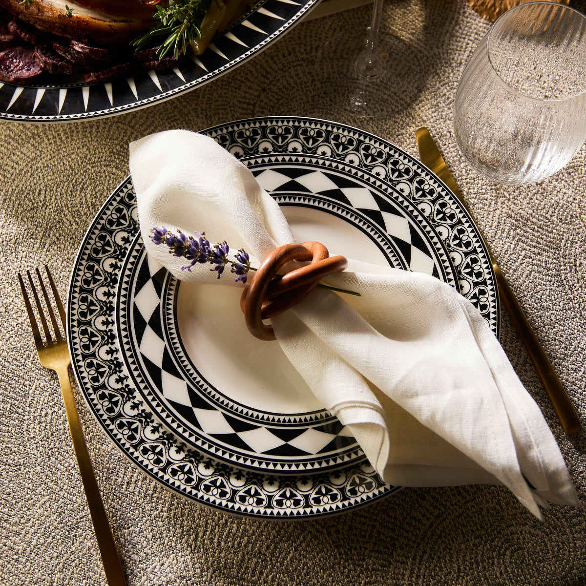 Sophisticated table setting featuring a Casablanca Rimmed Dinner Plate by Caskata Artisanal Home, complemented with gold cutlery and a beige napkin elegantly held by a decorative ring and lavender sprig, all set upon a textured tablecloth.
