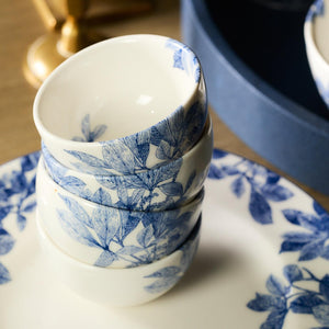 A stack of Arbor Snack Bowls by Caskata Artisanal Home, crafted from premium porcelain with a blue floral pattern, elegantly arranged on a matching plate atop a wooden surface. Each bowl is dishwasher safe, perfectly blending elegance with convenience.