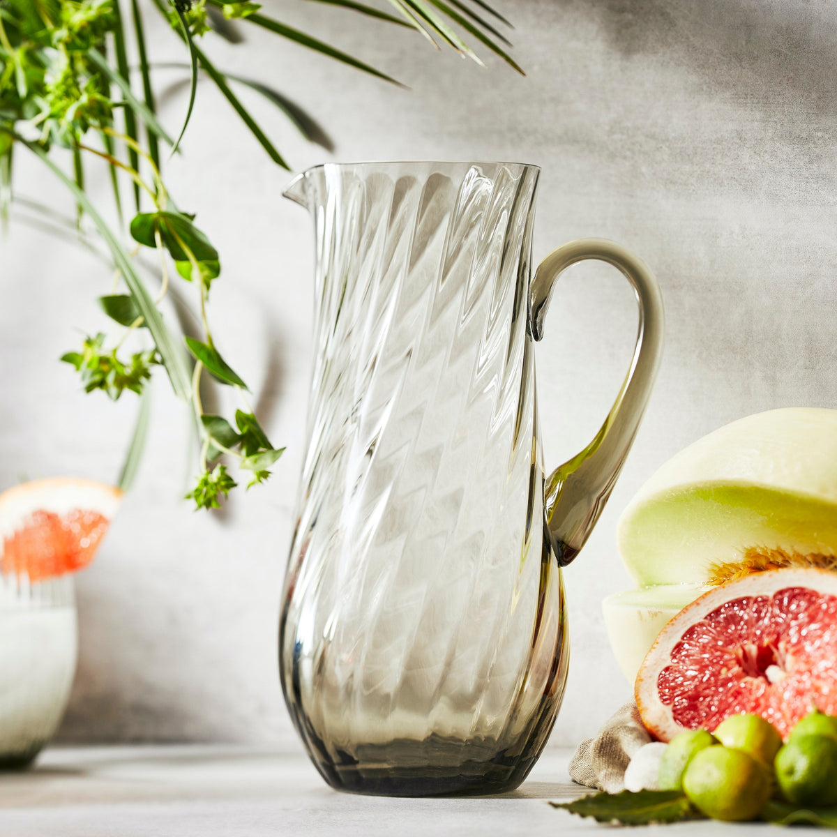 The mouth-blown crystal Quinn Smoke Pitcher by Caskata is shown on a sunny table with freshly cut citrus.