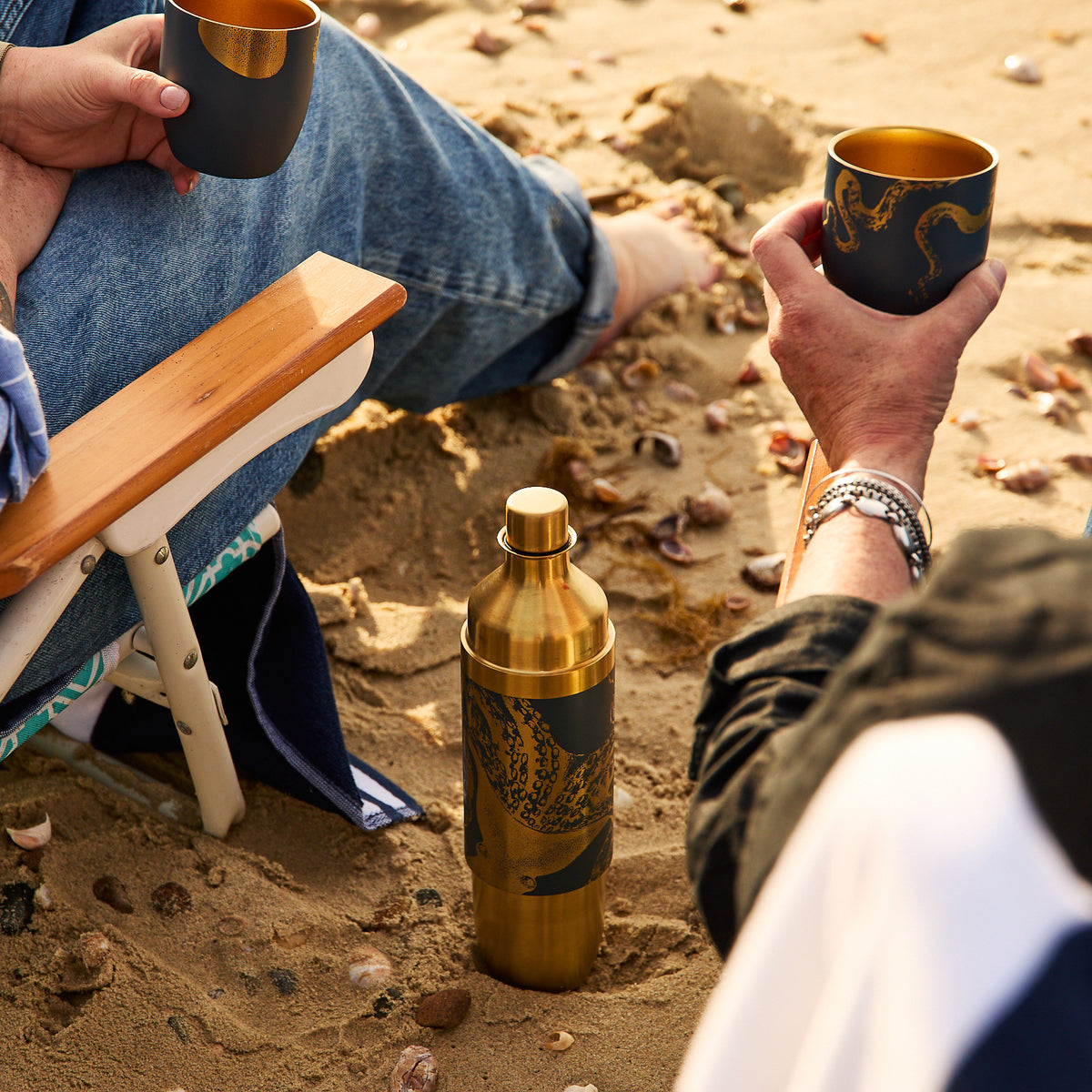 Two people on the beach hold matching cups, with a Caskata x High Camp Dark Blue Lucy Parkside Flask from Caskata resting in the sand between them.