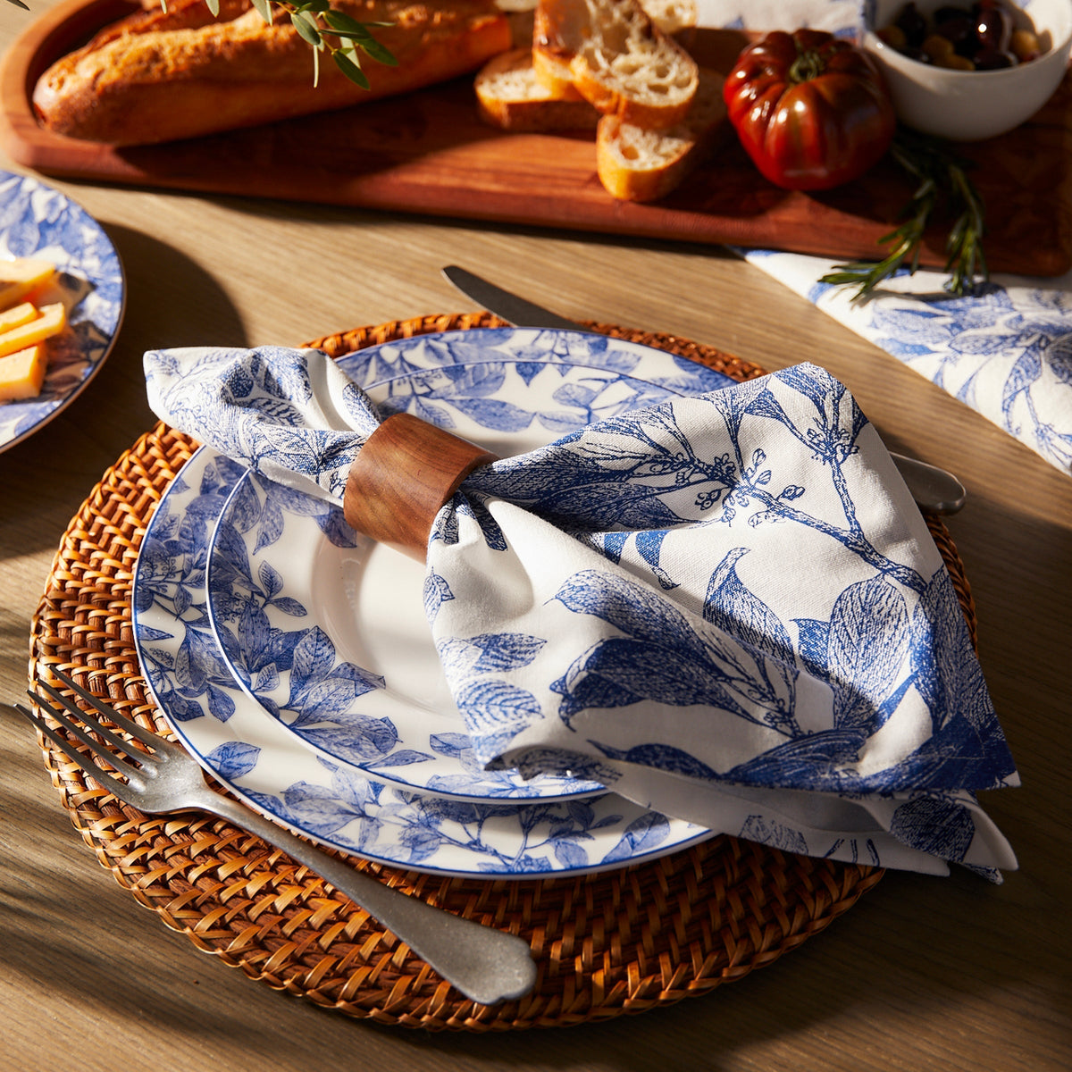 The table features a blue and white floral plate, an Arbor Birds napkin by Caskata in a wooden ring, cutlery, and a woven placemat. Bread and vegetables are on a wooden board behind. The stylish 100% cotton napkins are machine washable for easy care.