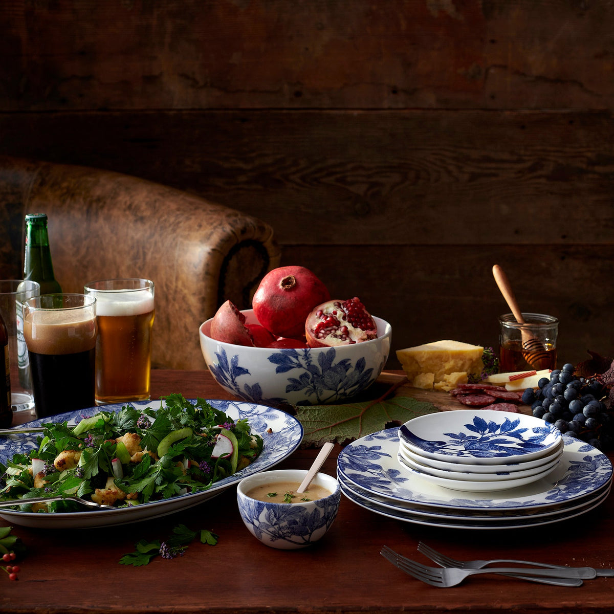 A rustic table setting with various foods, including salad, pomegranates, cheese, and drinks. Arbor Rimmed Dinner Plate by Caskata Artisanal Home, featuring blue floral patterns and delicate botanical details, are arranged around the table. The heirloom-quality dinnerware adds a timeless elegance to the scene.