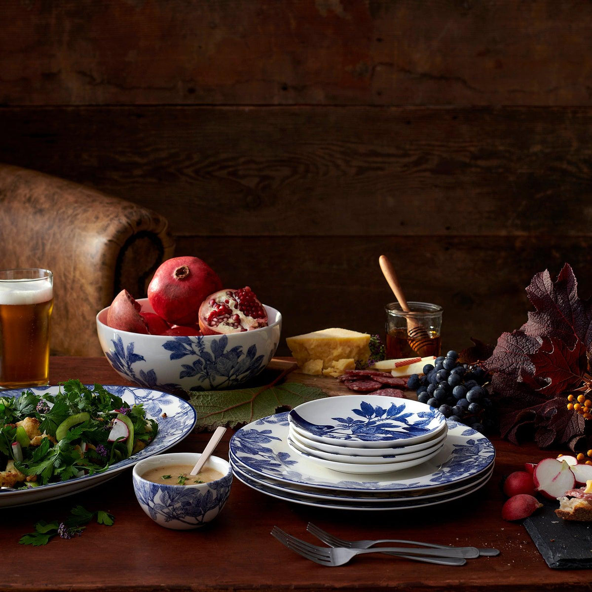 A rustic wooden table setting with Arbor Blue Birds Small Plates by Caskata Artisanal Home featuring botanical details, a salad, pomegranate, grapes, cheese, a beer, utensils, and various garnishes. This heirloom-quality dinnerware adds an elegant touch to any meal.