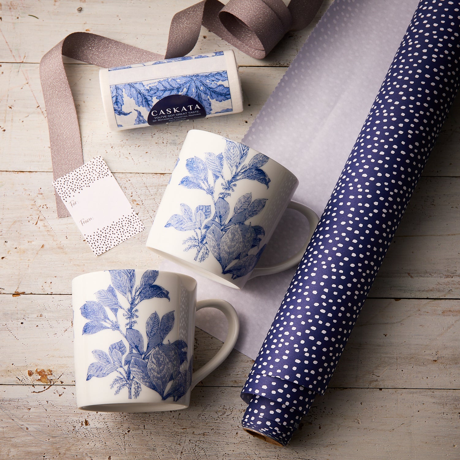 The "Arbor Coffee Talk" Bundle by Caskata features two blue botanical porcelain mugs, a purple polka-dot cocktail napkin roll, ribbon, and a Caskata box—beautifully arranged as the perfect gift for any coffee lover.