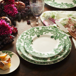 A table is set with Caskata Arbor Green Rimmed Dinner Plate, accompanied by a floral-patterned napkin, bread on a side plate, a glass, and a centerpiece of flowers and fruits.