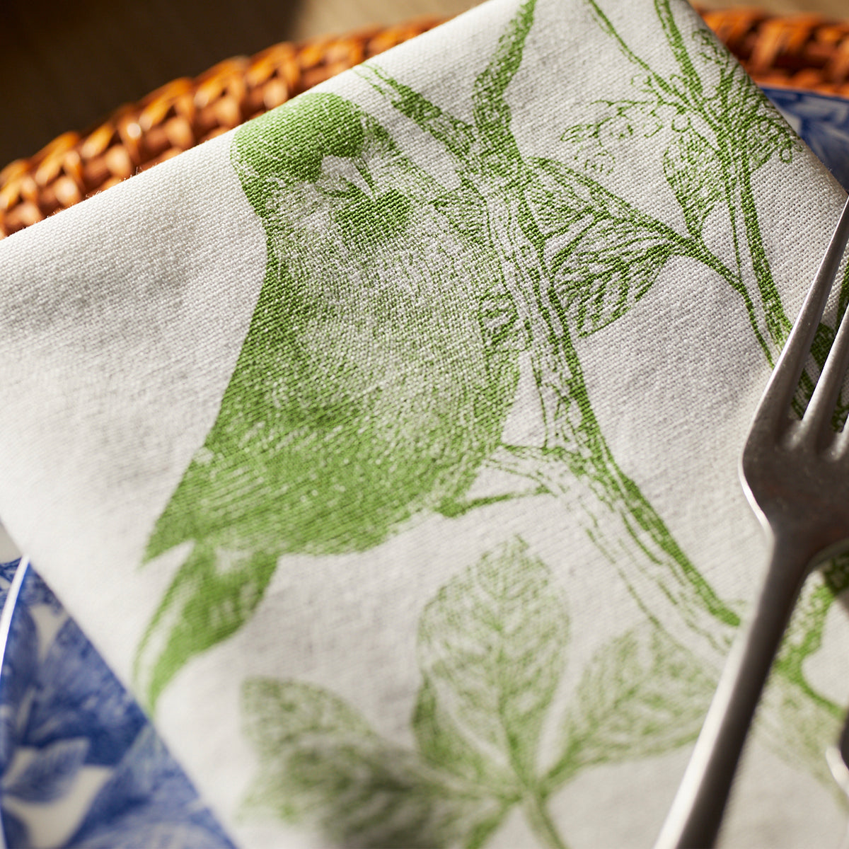 Part of Caskata&#39;s Greenhouse Luncheon Bundle, the white napkin with a green bird and branch design rests elegantly on a blue-patterned plate—ideal for simple gatherings. A fork nearby completes this refreshing spring table setting.