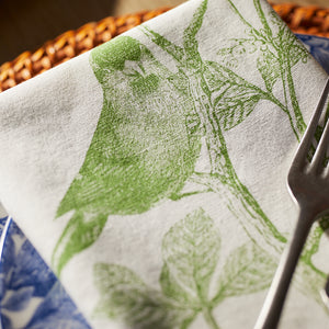 Part of Caskata's Greenhouse Luncheon Bundle, the white napkin with a green bird and branch design rests elegantly on a blue-patterned plate—ideal for simple gatherings. A fork nearby completes this refreshing spring table setting.