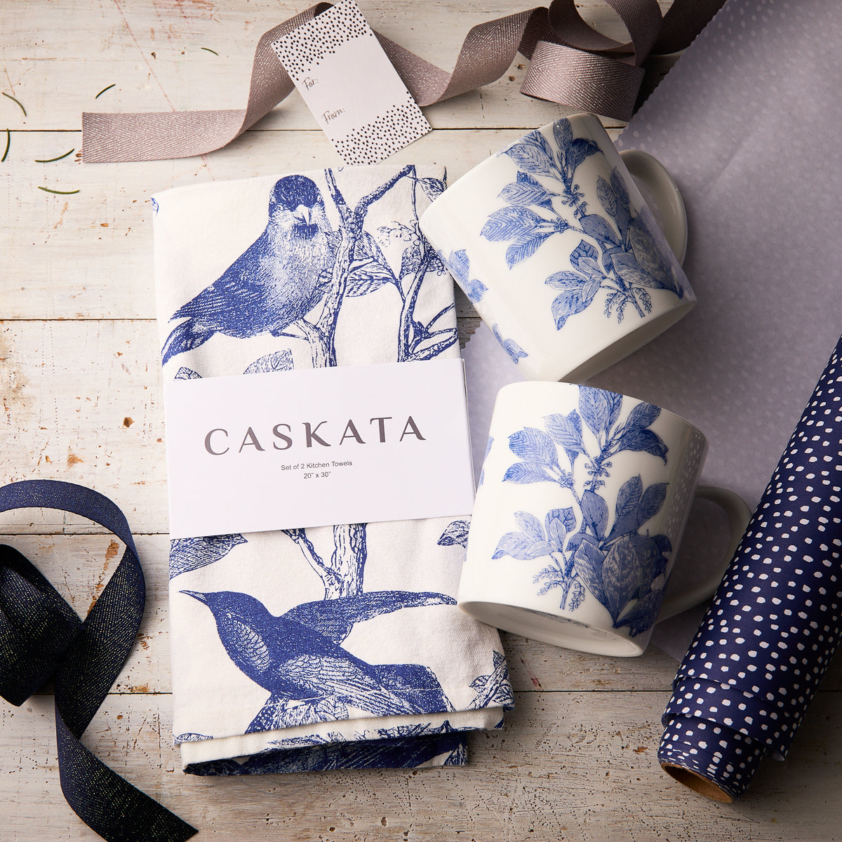 The "Arbor In the Kitchen" Bundle by Caskata features two blue botanical porcelain mugs and themed kitchen towels, beautifully arranged with ribbon and wrapping paper for a charming gift set.
