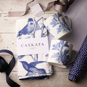 The "Arbor In the Kitchen" Bundle by Caskata features two blue botanical porcelain mugs and themed kitchen towels, beautifully arranged with ribbon and wrapping paper for a charming gift set.