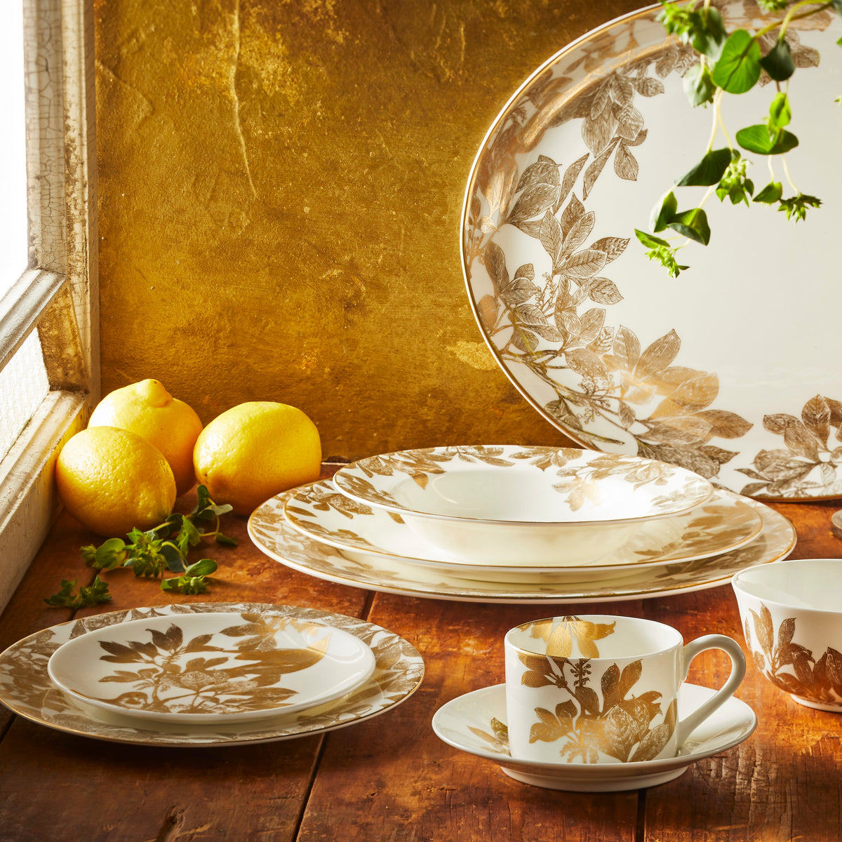 Warm sunlight shines through a window on Caskata&#39;s Gold Arbor Collection. Products featured include the canapé plate, salad plate, cup and saucer, charger, dinner plate, rimmed soup bowl, small bowl and platter.