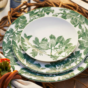A set of three Caskata Arbor Green Small Plates with green leaf patterns is neatly stacked, placed on a braided charger. A small orange flower is partly visible in the foreground, adding a touch of color to the botanical dinnerware. These plates are also dishwasher safe for easy cleanup.