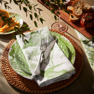 A table setting with a green-patterned plate on a rattan charger, complemented by Caskata Arbor Birds Verde Dinner Napkins and silver cutlery, surrounded by bread, cheese, and a glass of wine.
