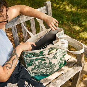 Seated on a wooden bench, a person places their laptop into the Caskata x Steele Everyday Tote Bag Arbor Green by Caskata, handcrafted in the USA, alongside a rolled-up newspaper and other essentials.