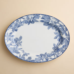 The Arbor Oval Rimmed Platter by Caskata Artisanal Home features a large, white porcelain design with an elegant blue botanical leaf pattern around the rim, set against a beige background.