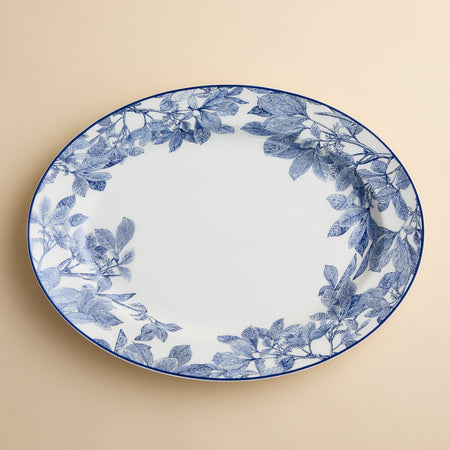 The Arbor Oval Rimmed Platter by Caskata Artisanal Home features a large, white porcelain design with an elegant blue botanical leaf pattern around the rim, set against a beige background.