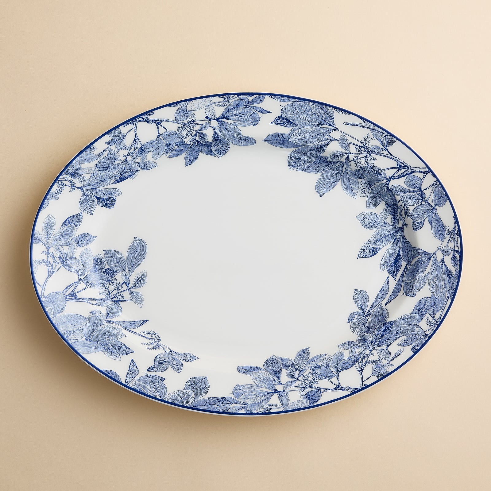 The Arbor Oval Rimmed Platter by Caskata Artisanal Home features a large, white porcelain design with an elegant blue botanical leaf pattern around the rim, set against a beige background.