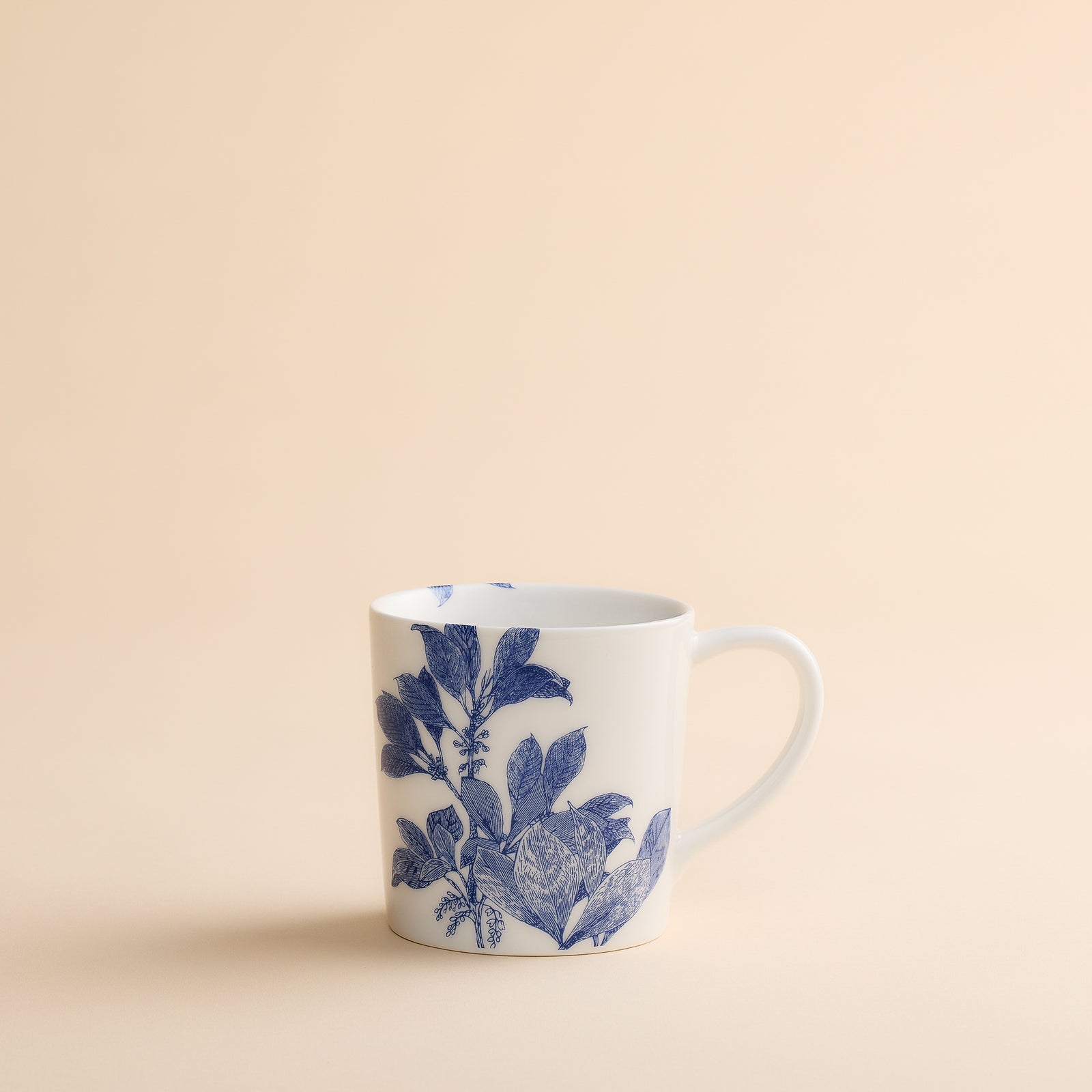 The Arbor Mug by Caskata Artisanal Home showcases a white porcelain body adorned with a blue botanical pattern, displayed on a plain beige background.