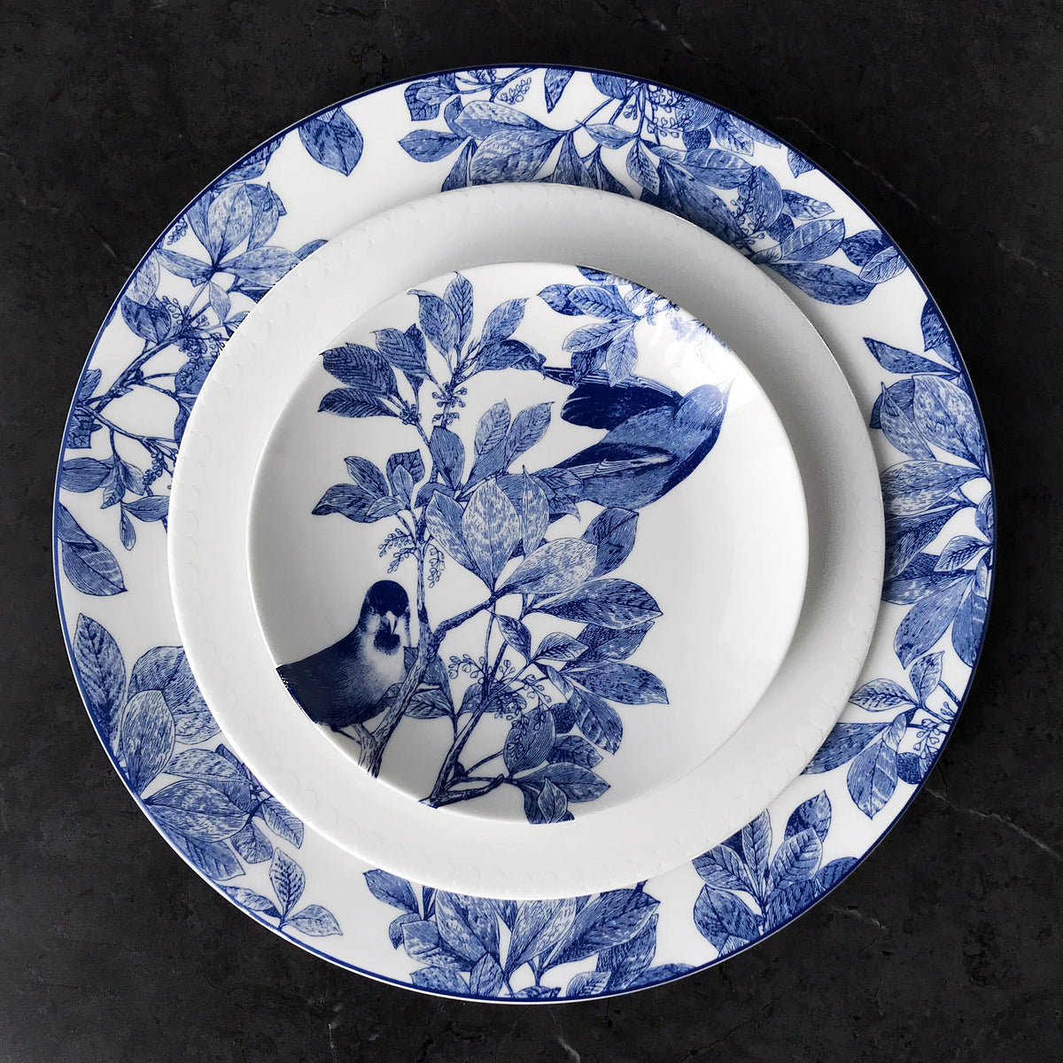 Two plates on a dark surface. The smaller plate is atop the larger one, both featuring a blue and white design with birds and botanical details. The plates are Arbor Blue Birds Small Plates by Caskata Artisanal Home.