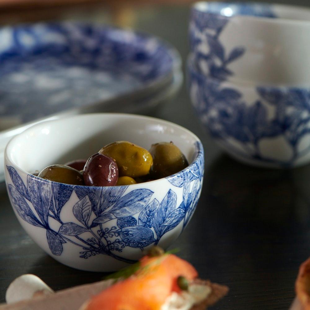 A small porcelain bowl adorned with blue floral and leafy branch patterns on a white background, both dishwasher and microwave safe. The Arbor Snack Bowl by Caskata Artisanal Home.