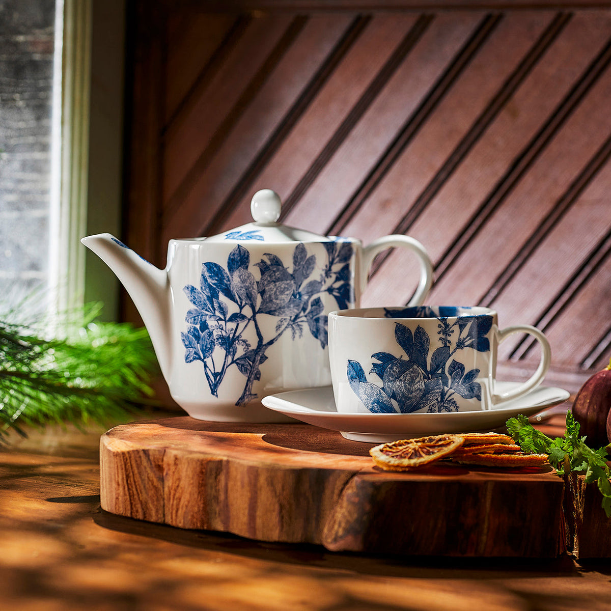 Arbor blue and white bone china teapot and tea cup and saucer from Caskata is shown on a rustic board in a window.