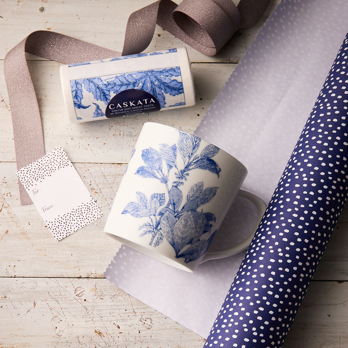 The "Arbor with Breakfast" Bundle by Caskata features a blue botanical porcelain mug, a matching box, polka dot wrapping paper, ribbon, and a gift tag—perfectly arranged as a charming gift set on a wooden surface.