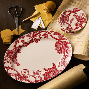 Caskata’s “Happy Hosting” Bundle features oval and round Arcadia Crimson plates with red floral designs, styled with a yellow ribbon, scissors, pen, gift tags, and an oversized platter for a festive presentation.