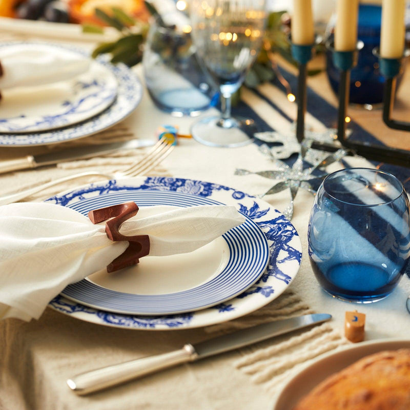 A dining table is set with blue and white Newport Stripe Rimmed Salad Plates by Caskata Artisanal Home, high-fired porcelain salad plates, blue glassware, and a white napkin held by a wooden napkin ring. A bread loaf, candles, and decorative stars are also on the table.