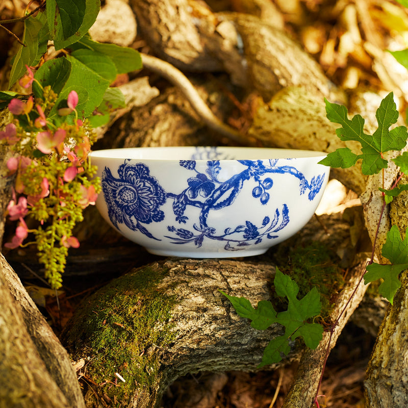 The Caskata Arcadia Serving Set features a white ceramic bowl with blue floral patterns, elegantly displayed on tree roots amid green leaves and pink flowers.