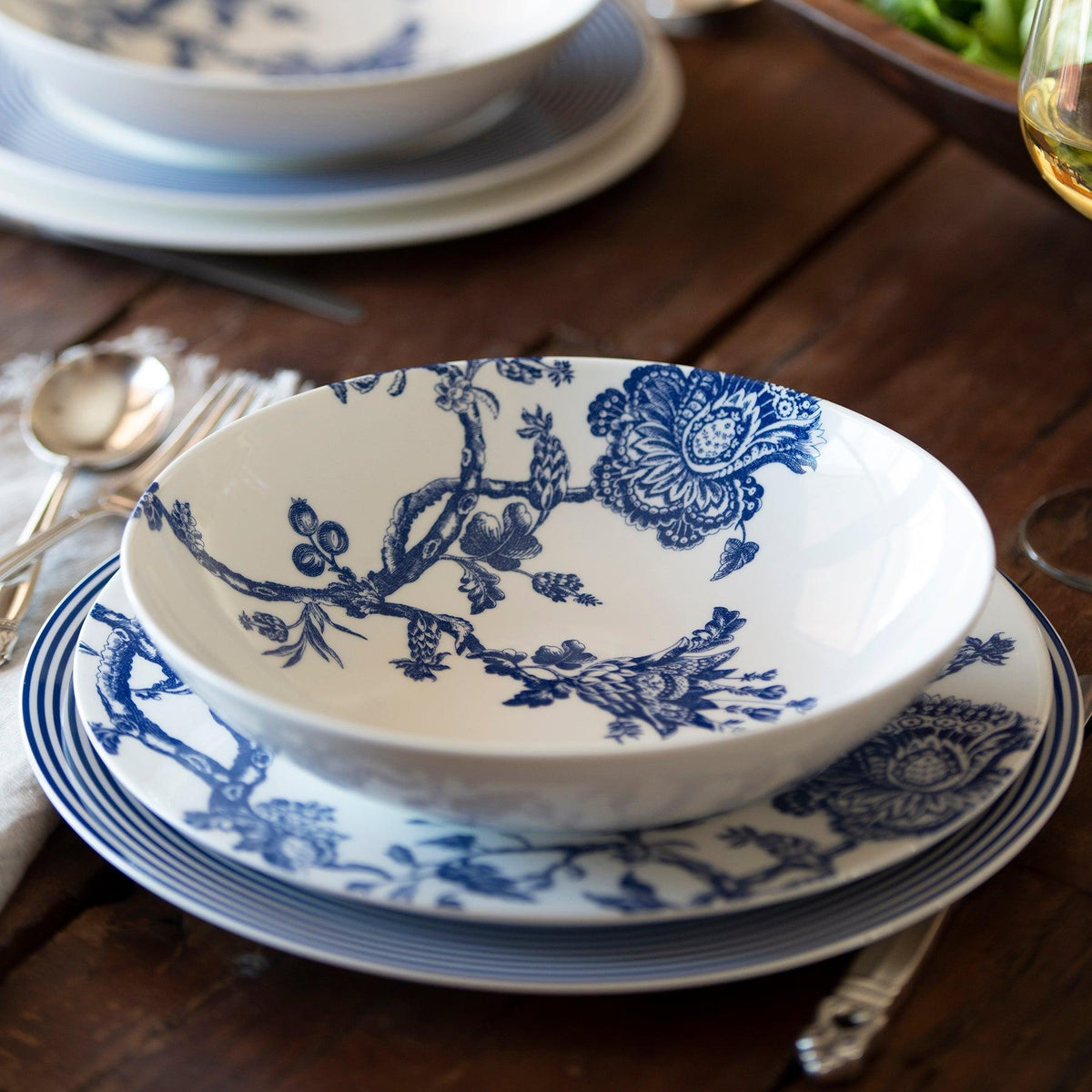 A table set with a generous Caskata Artisanal Home Arcadia Entrée Bowl, matching plate, fork, spoon, and knife on a wooden surface. A glass of white wine and a green salad are in the background. This elegant scene features premium porcelain dinnerware from the Williamsburg Foundation collection.