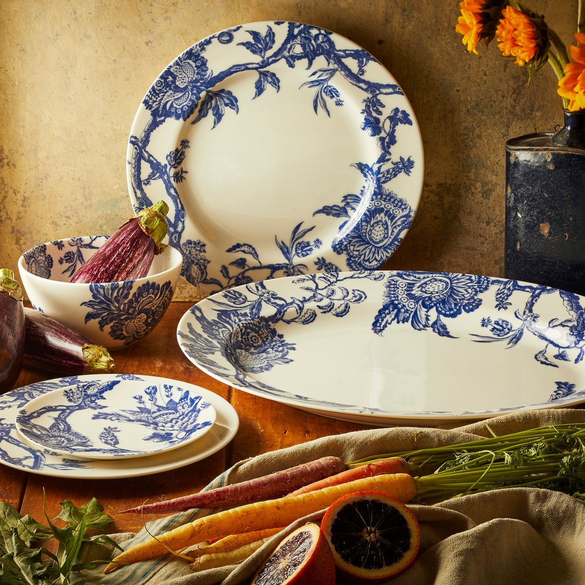 The blue Arcadia Collection by Caskata is displayed on a moody tabletop with fresh vegetables and flowers. The large oval platter, tall cereal bowl, dinner, salad and canapé plates are displayed.