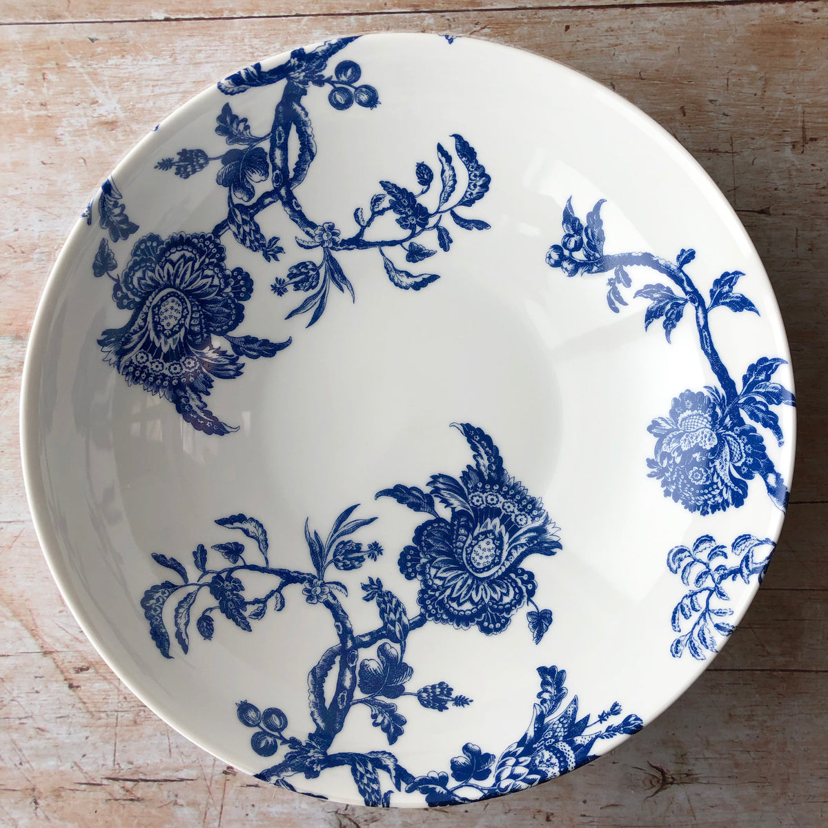 An Arcadia Wide Serving Bowl by Caskata Artisanal Home, featuring a blue and white floral design, placed on a wooden table with dimensions.