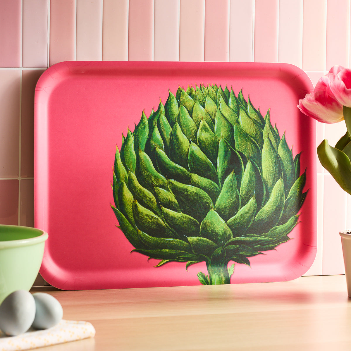 The Artichoke Large Rectangular Birchwood Tray by Caskata, featuring an artichoke design, leans against a pink-tiled wall while a tulip, green bowl, blue eggs, and folded cloth adorn the surface.
