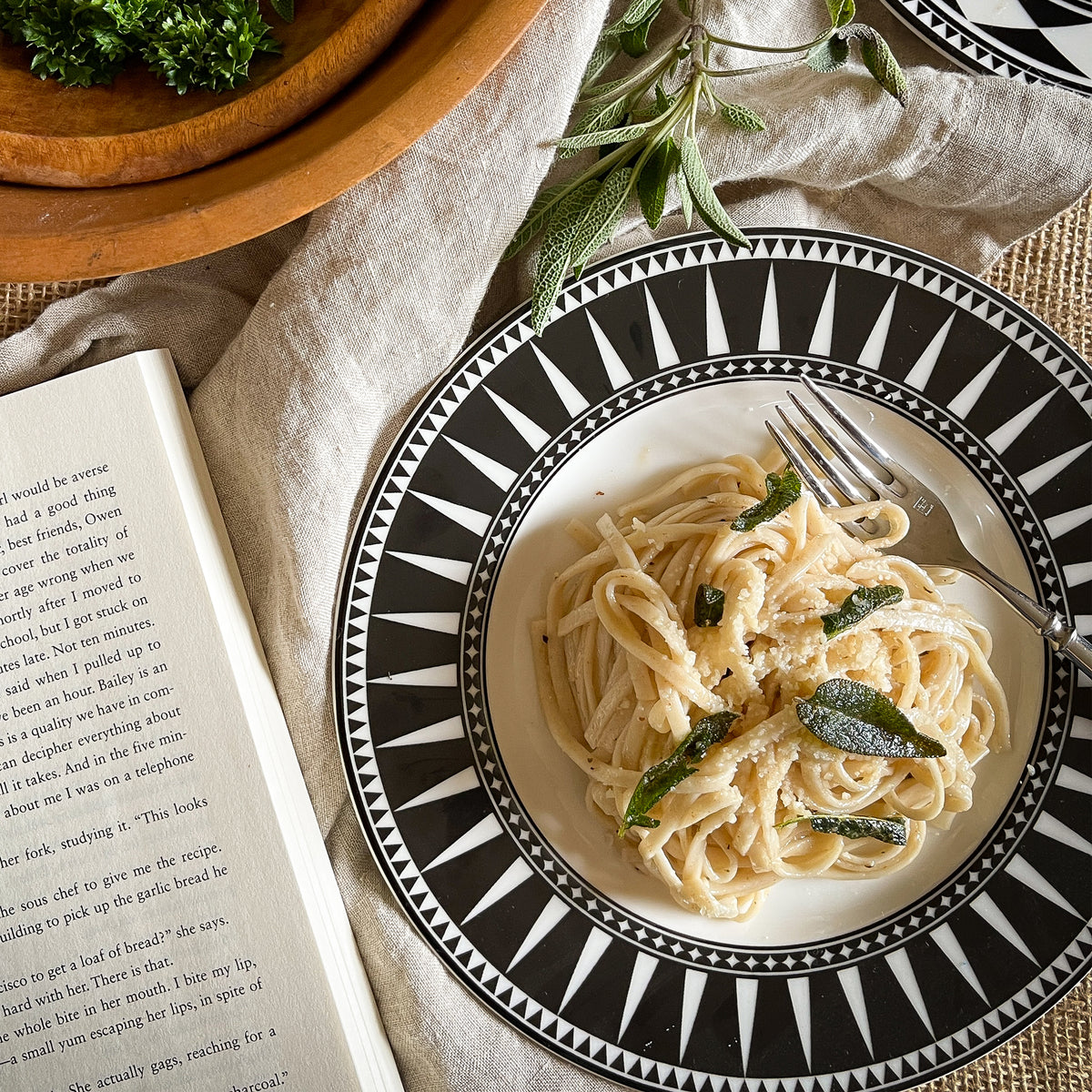 A plate of spaghetti with sage and cheese is served on the Caskata Marrakech Ultimate Set, placed beside a fork, an open book, and a bowl of greens atop a linen tablecloth.