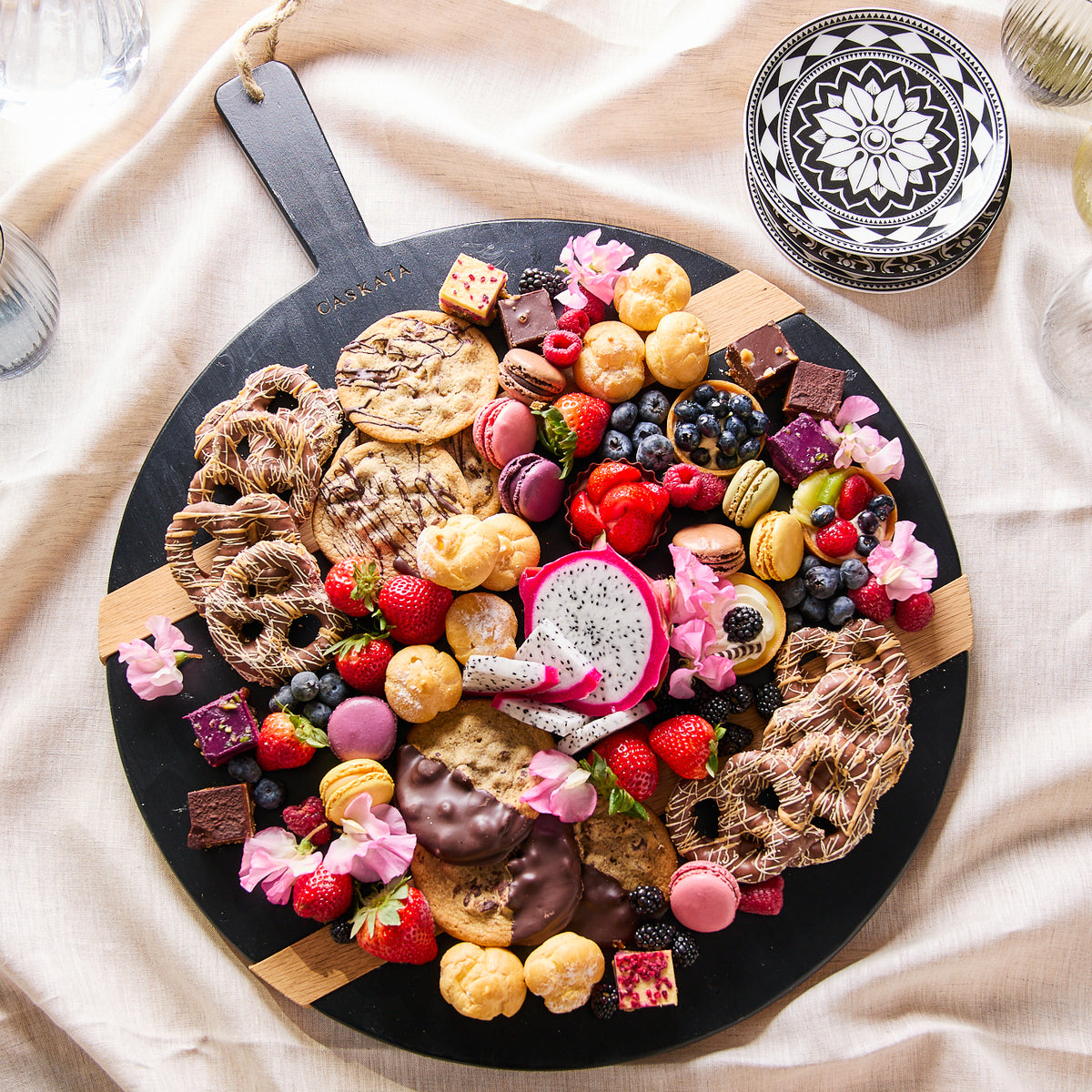 A vibrant charcuterie board with a variety of fruits, chocolates, and pastries on a Black &amp; Natural Round Charcuterie Big Board made from reclaimed European timbers by Etu Home, garnished with edible flowers.