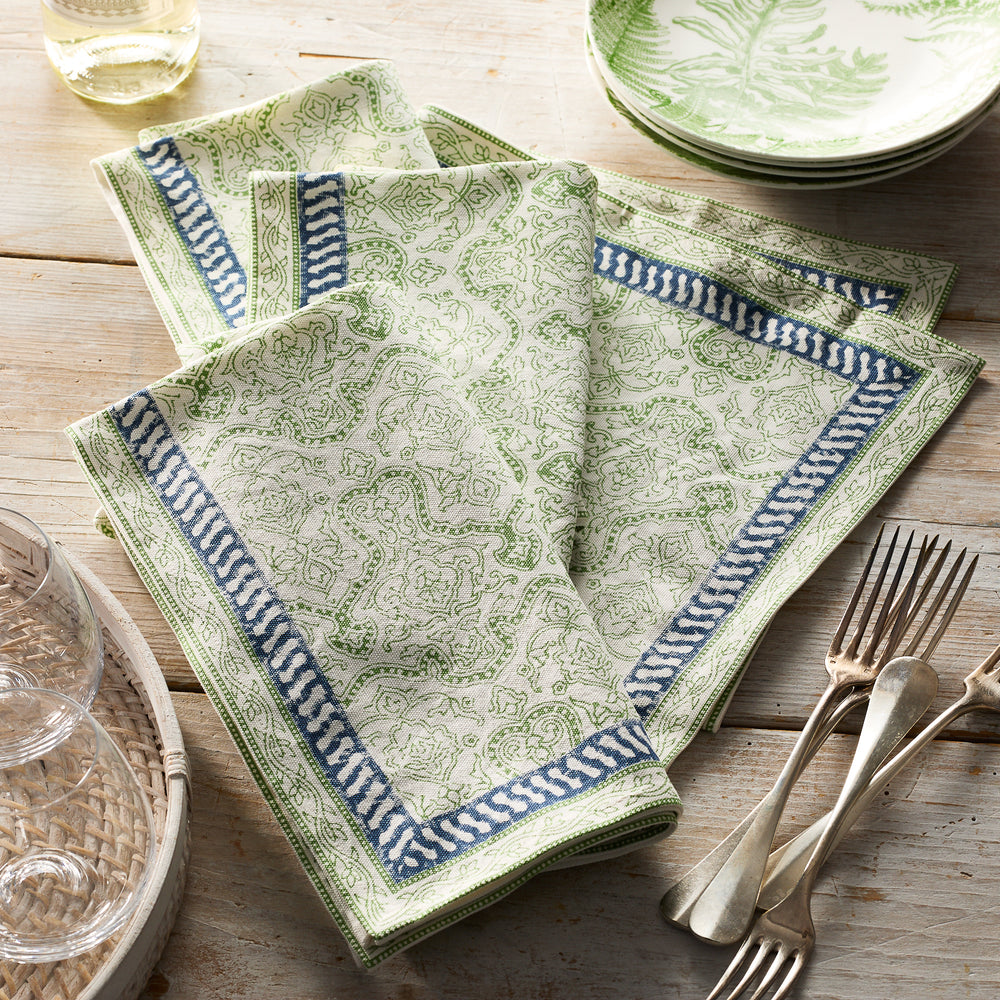 Caskata’s Block Island Block Print Dinner Napkins in green and white with blue borders bring bohemian flair to a wooden table, placed next to stacked plates, glassware, and cutlery.
