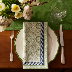 A table setting with a green-bordered plate, Caskata Block Island Block Print Dinner Napkins, fork, knife, wine glass, and a green table runner with floral decor for a bohemian look.