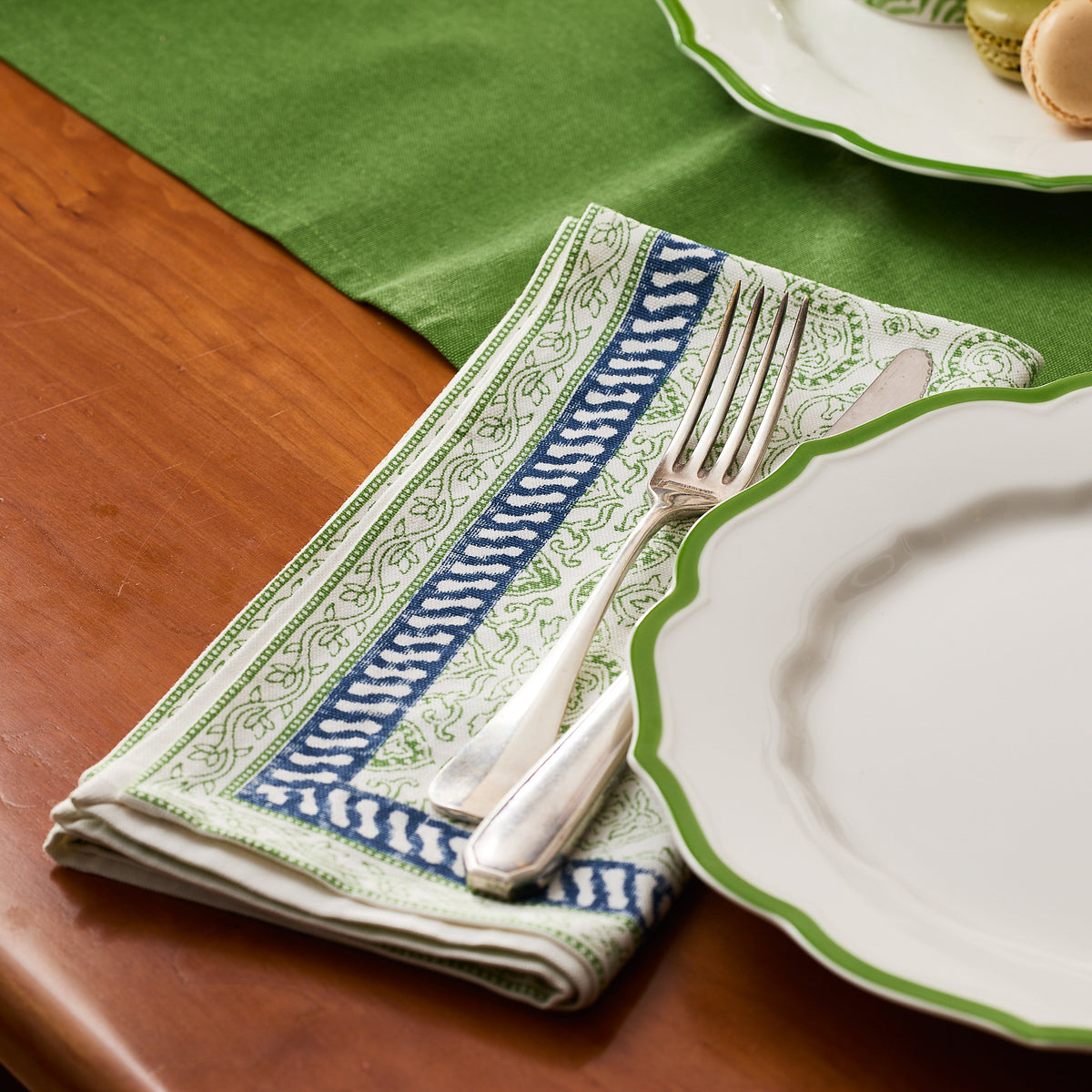 A folded Caskata Block Island Block Print Dinner Napkin with a fork and knife sits beside a white plate with green rim on a wooden table and green runner, adding boho charm to your table décor.