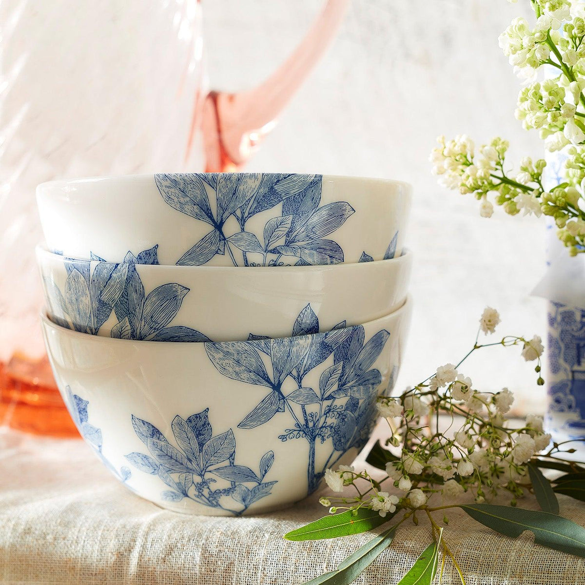 Three premium Caskata Arbor Cereal Bowls with blue botanical patterns are stacked together, alongside a bouquet of small white flowers and a clear pink pitcher in the background.