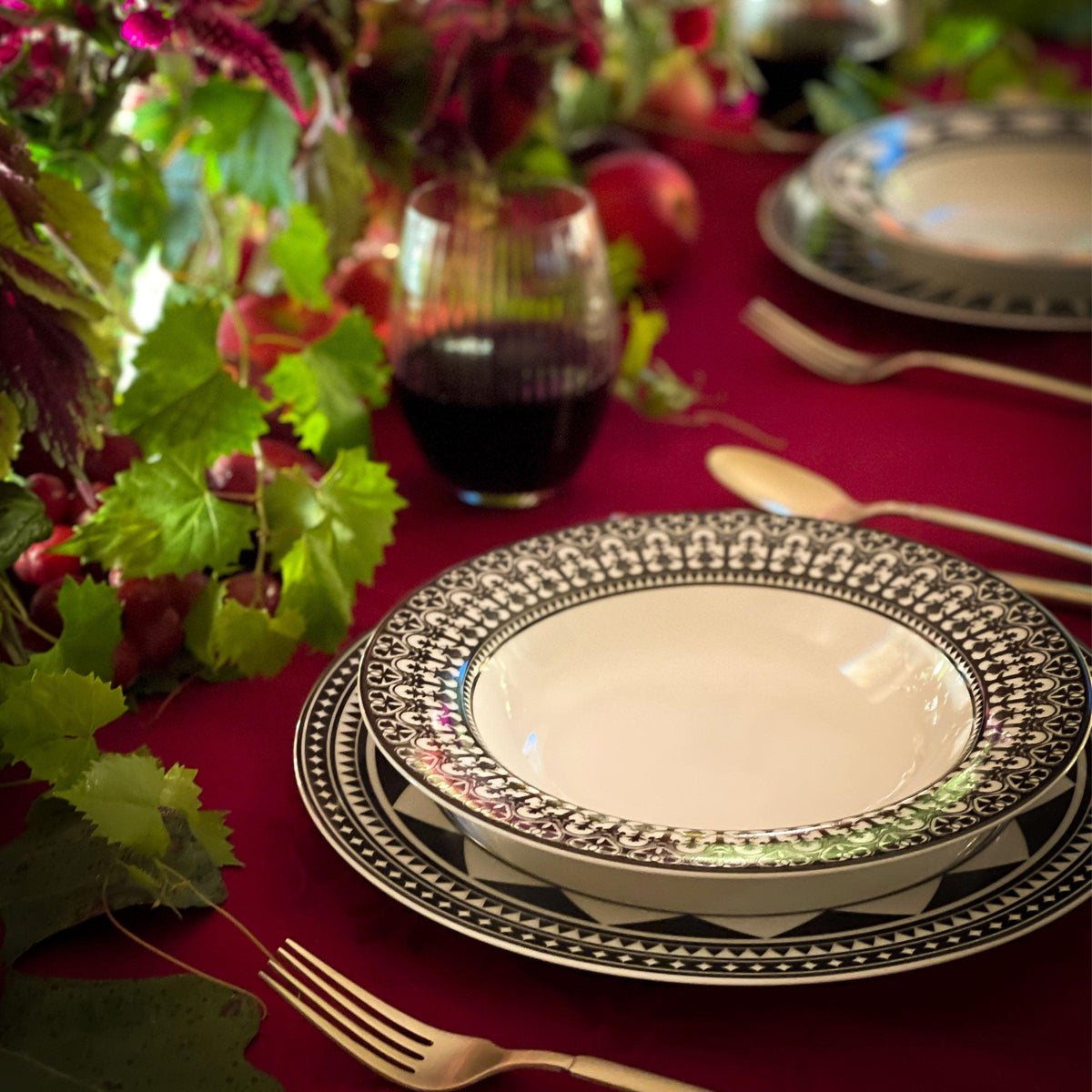 Elegant dining table setting with ornate Casablanca rimmed soup bowls from the Caskata Artisanal Home Geometrics Collection and decorative foliage on a red tablecloth.