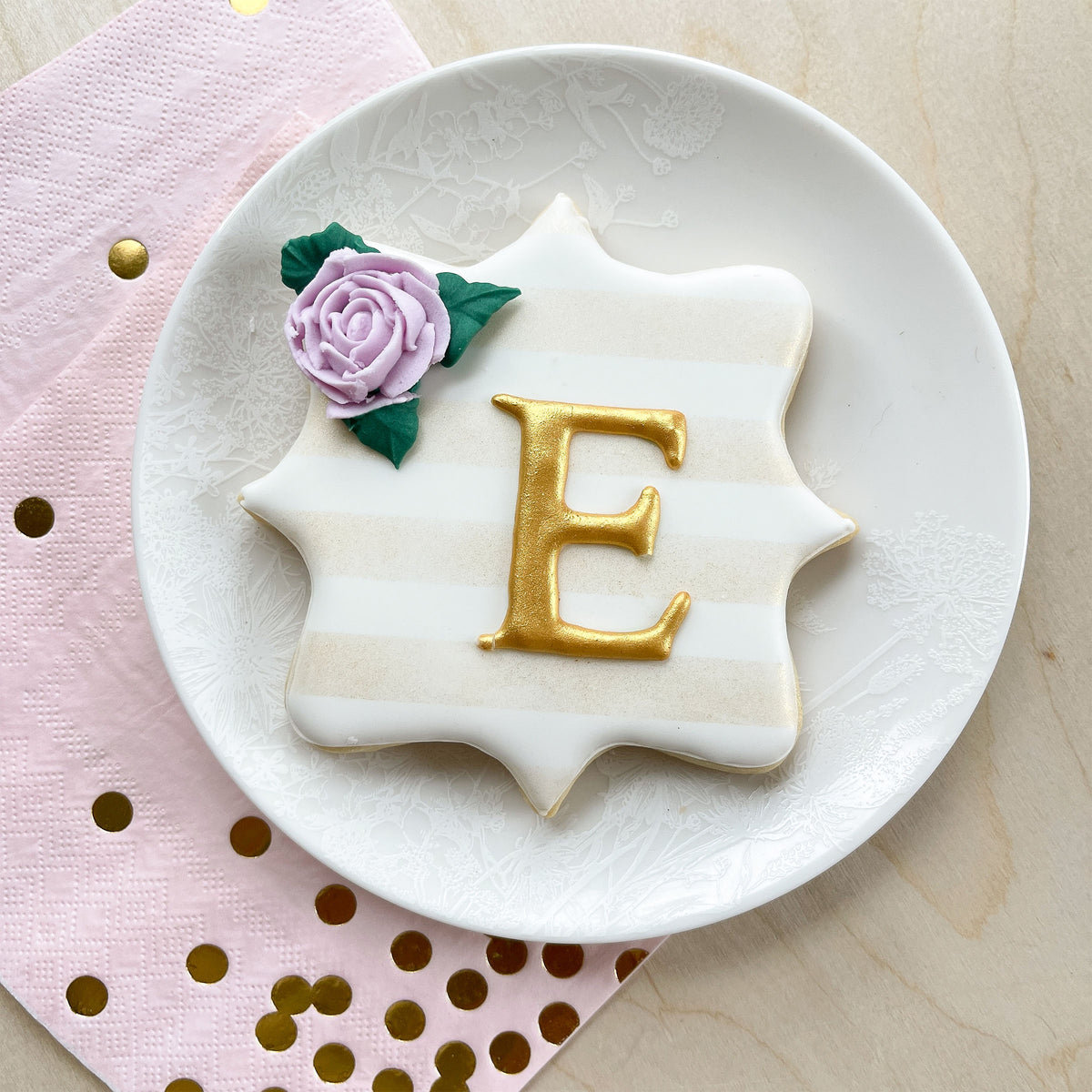 A decorative sugar cookie with a gold &quot;E&quot; and purple flower rests on a Caskata Summer White Small Plate, set atop a pink napkin with gold dots.