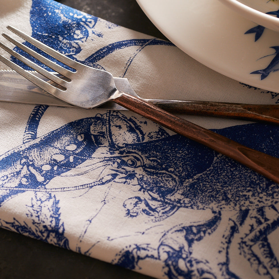 A fork and knife rest on a Caskata Blue Lobster Dinner Napkin next to a white plate with a blue floral pattern.