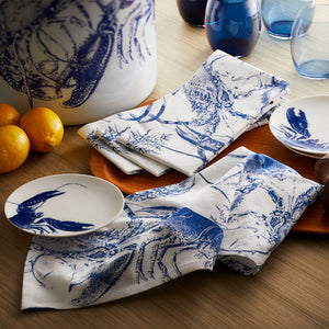 A charming table setting showcases blue crab-printed plates and Caskata's Blue Lobster Dinner Napkins, made of 100% cotton, on a wooden surface. It is accompanied by whole oranges and blue glasses.