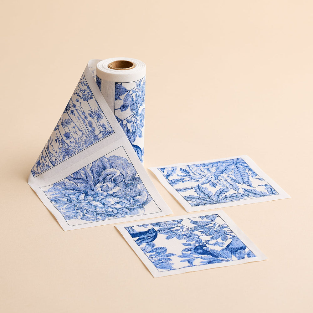 A roll of MY DRAP’s Botanicals Cocktail Napkin Roll is unspooled, with three individual square napkins featuring blue and white floral and botanical designs placed beside it on a beige surface—evoking the charming motifs often found on handcrafted cotton napkins.