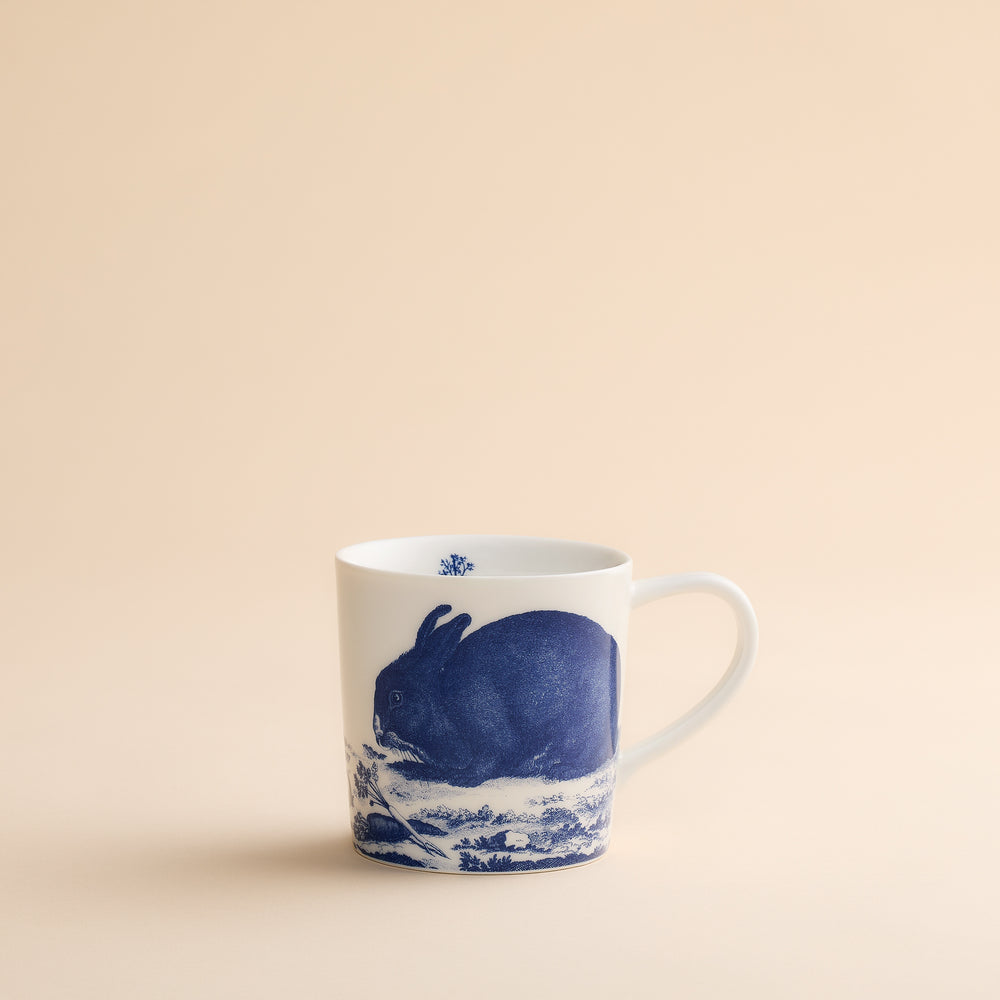 The Caskata Artisanal Home Bunnies Mug is a white porcelain mug featuring a charming blue rabbit illustration on the side, set against a simple beige background. This delightful Bunnies Mug adds a playful touch to your spring mornings and is perfect for enjoying your favorite beverages.