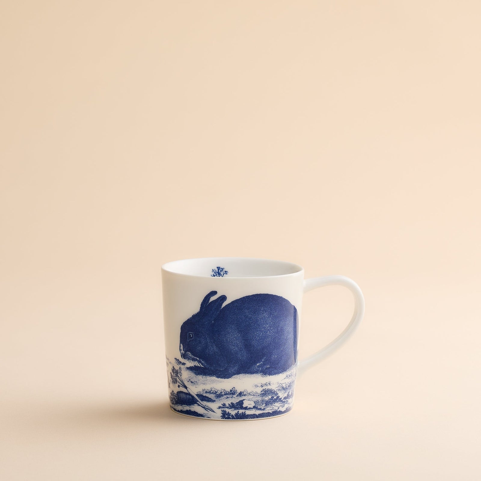The Caskata Artisanal Home Bunnies Mug is a white porcelain mug featuring a charming blue rabbit illustration on the side, set against a simple beige background. This delightful Bunnies Mug adds a playful touch to your spring mornings and is perfect for enjoying your favorite beverages.