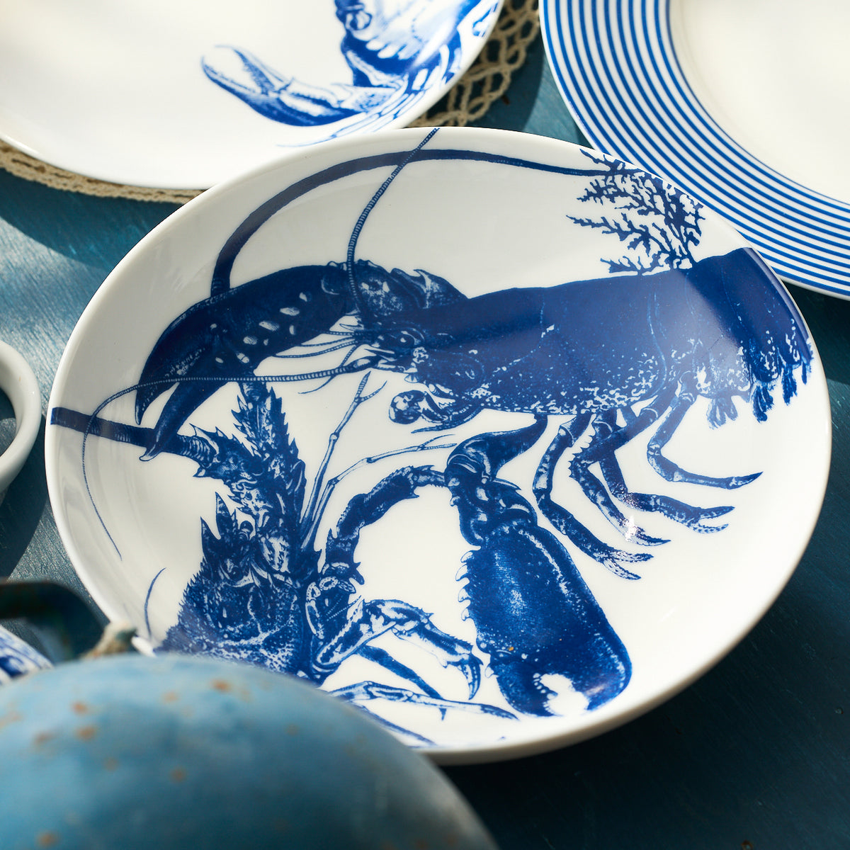 The Caskata Lobster Entrée Bowl, a white ceramic bowl with a detailed blue lobster illustration, is displayed alongside other blue and white dishware.