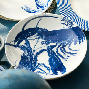 The Caskata Lobster Entrée Bowl, a white ceramic bowl with a detailed blue lobster illustration, is displayed alongside other blue and white dishware.